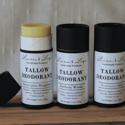 three Linen and Logs deodorants inblack biodegradable tubes with white labels sitting on a multibar slat holder. Far left deodorant has lid off and leaning on the side. and the deodorant is sticking up. The other two deodorants have their lids on.